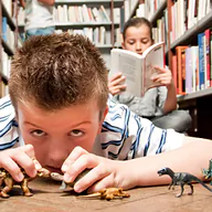 Two children are in a library. One child is lying on the floor, closely examining small toy dinosaurs and other animals. He has short brown hair and is wearing a striped shirt. The other child is sitting on the floor, reading a book with her hair tied back. In the background, there are bookshelves filled with books.