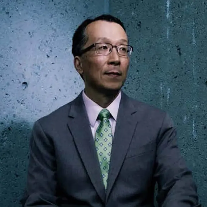 A man wearing glasses sits in front of a textured blue wall. He has short black hair and wears a light pink shirt with a green tie that has a diamond pattern. He is dressed in a dark gray suit and looks slightly to the side with a calm expression.