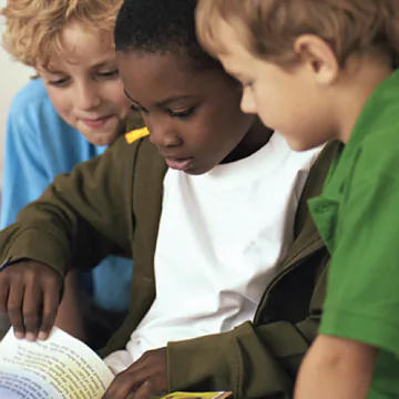 Three children are gathered together, focused on a book. One child, wearing a green shirt, sits in front, closely inspecting the pages. The child on the left, with curly blonde hair, leans in to see better, while another child, who has darker skin and is wearing a dark green jacket, looks thoughtfully at the book. The atmosphere is collaborative and engaging, suggesting they are sharing ideas or stories from the book.